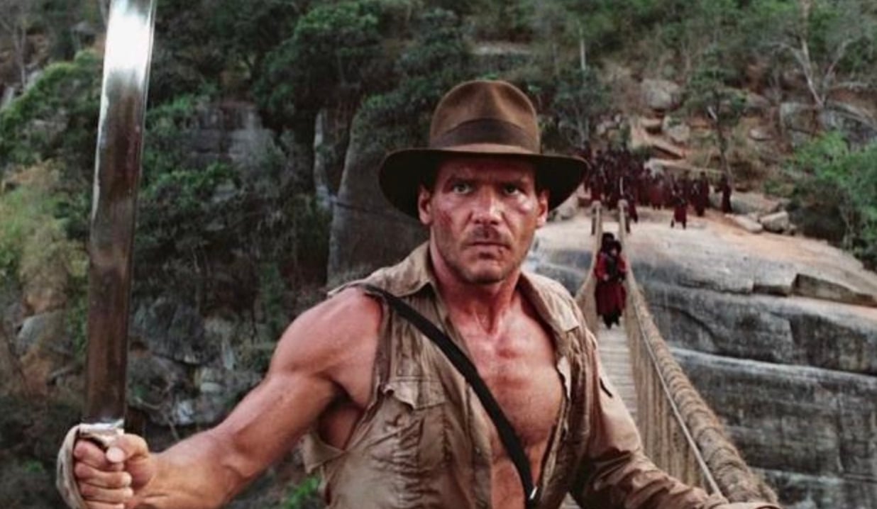A man in a brown fedora and ripped shirt holds a sword, standing on a rope bridge with dense forest and people in the background, looking intense and ready for action.
