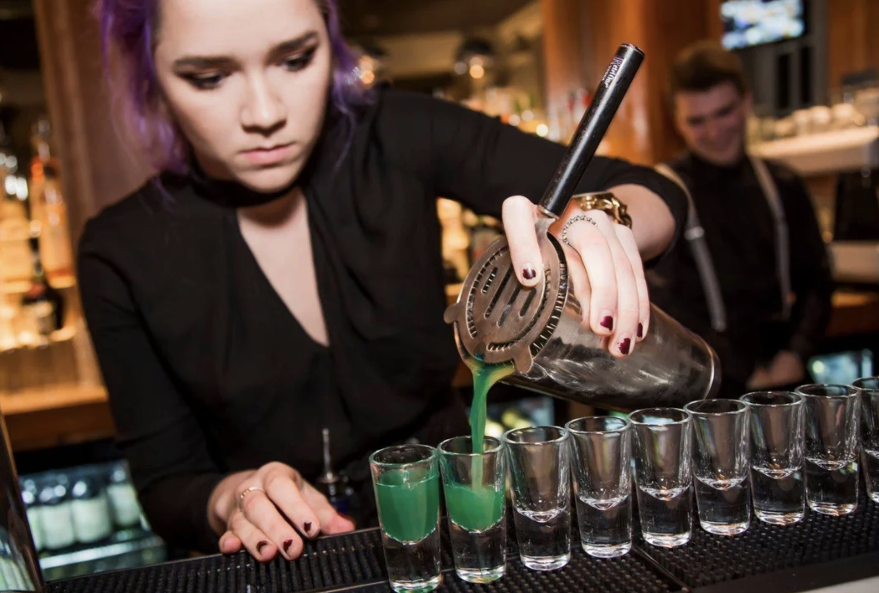 A bartender with purple hair pours a green cocktail from a shaker through a strainer into a row of shot glasses at a bar, while a man in the background watches and smiles.