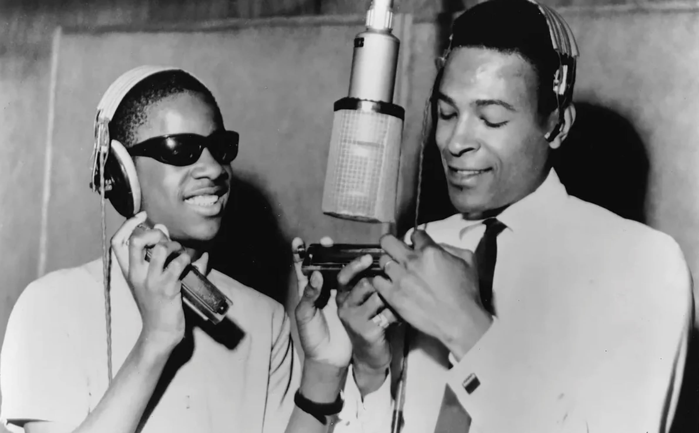 Two Black men stand in a recording studio. One wears sunglasses and holds a harmonica, smiling, while the other, dressed in a shirt and tie with headphones, looks at the harmonica under a large microphone.
