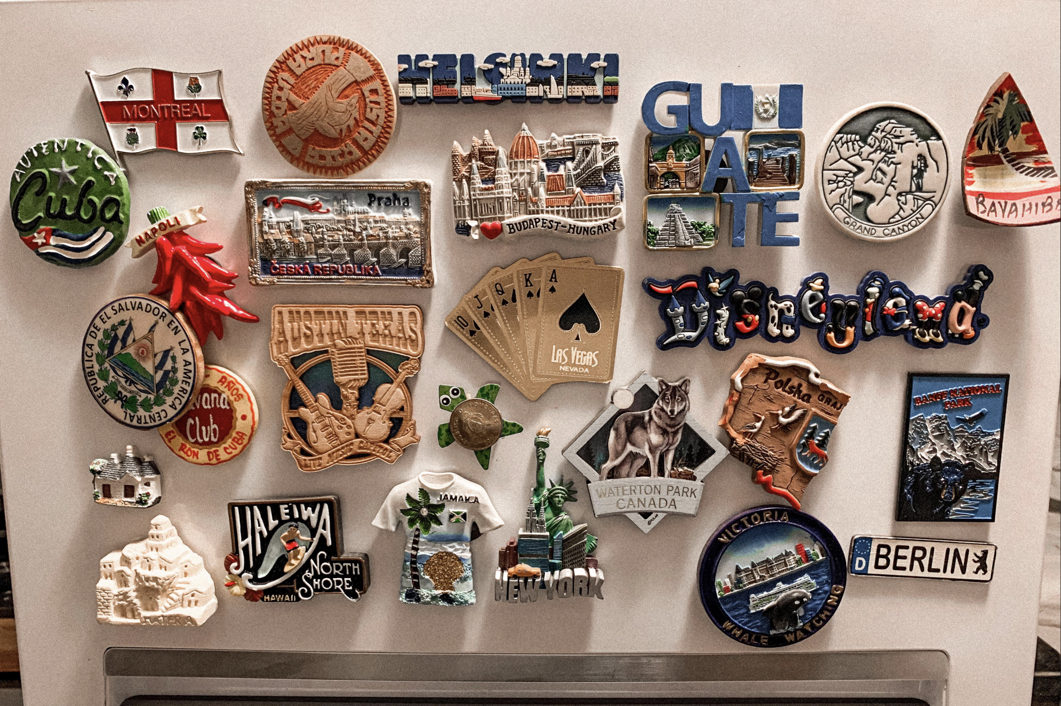 A collection of colorful souvenir magnets from various cities and countries, including Montreal, Cuba, Prague, Berlin, Las Vegas, Budapest, Banff, and more, displayed on a metallic surface.