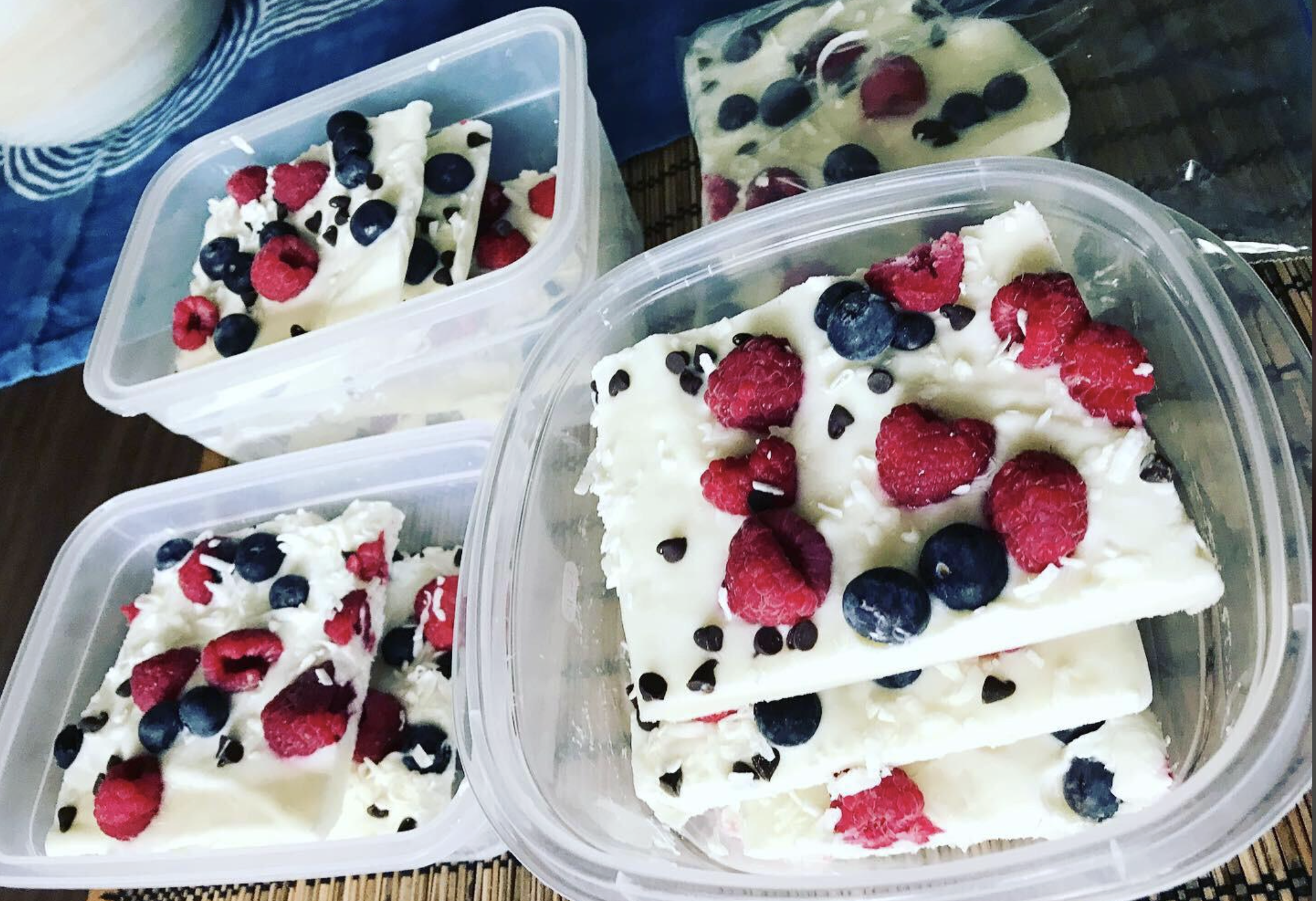 Rectangular slices of white frozen yogurt bark topped with raspberries, blueberries, and mini chocolate chips, neatly stacked in clear plastic containers on a table.