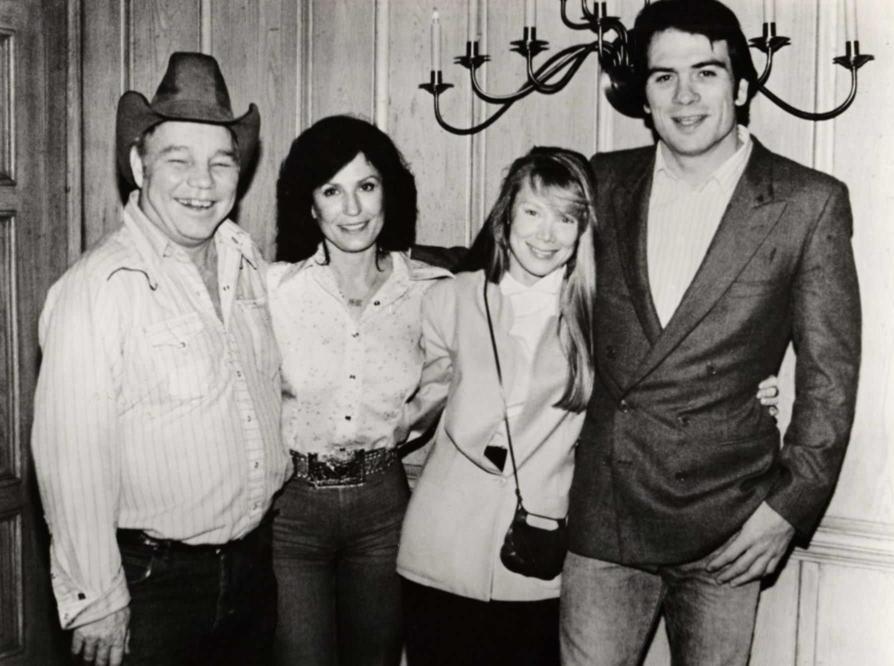 A black-and-white photo of two men and two women standing together and smiling indoors. One man wears a cowboy hat and Western shirt; the others wear jackets. A candle-style wall fixture is visible behind them.