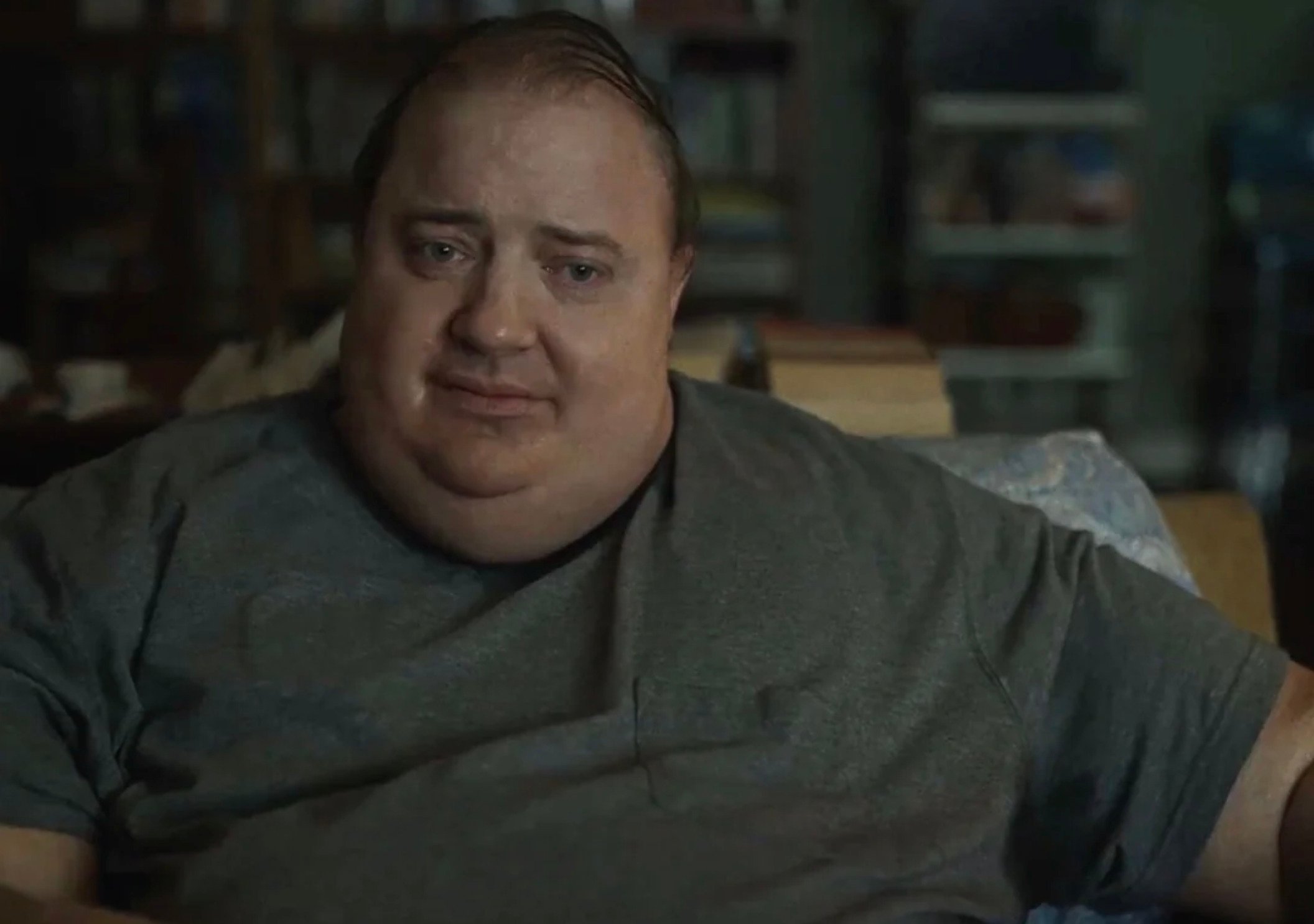 A man with a sad expression sits on a couch, wearing a gray T-shirt, with cluttered shelves in the blurry background.