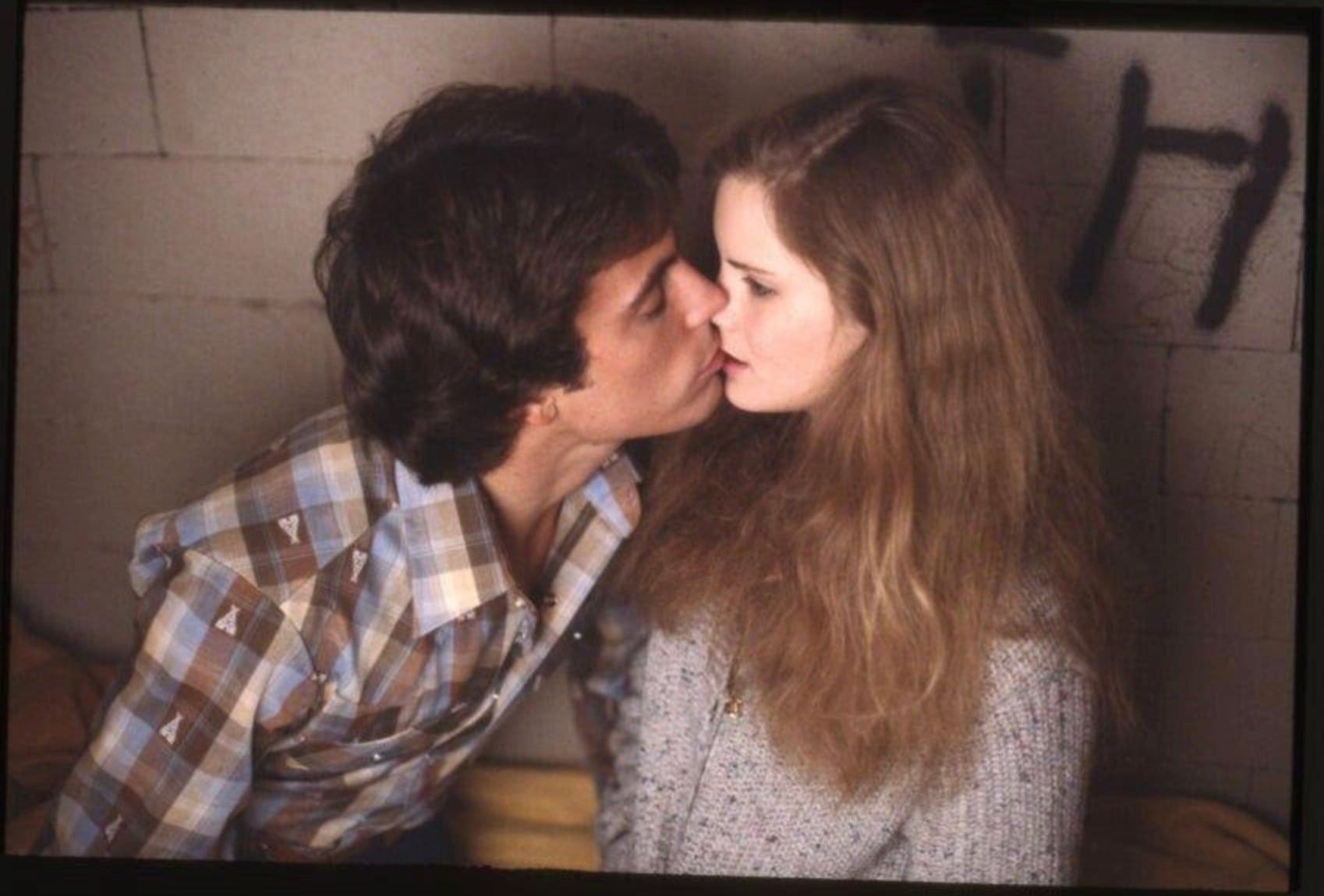 A young man in a plaid shirt leans in closely, appearing about to kiss a young woman with long, wavy hair who looks ahead with a serious expression. They are sitting against a graffiti-marked brick wall.