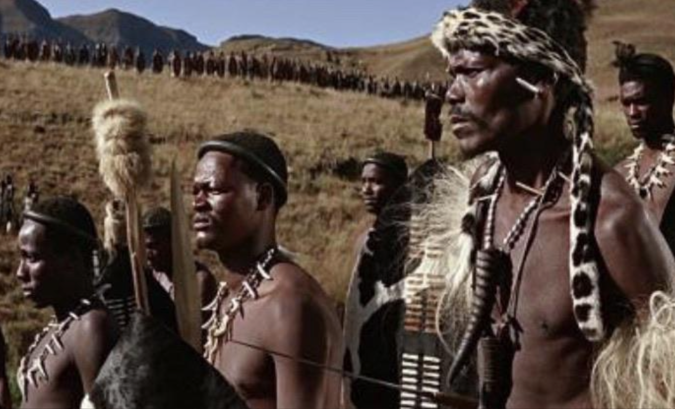 A group of men in traditional Zulu attire stand outdoors in a grassy landscape, holding shields and spears, with more people visible in a long line on a distant hillside.