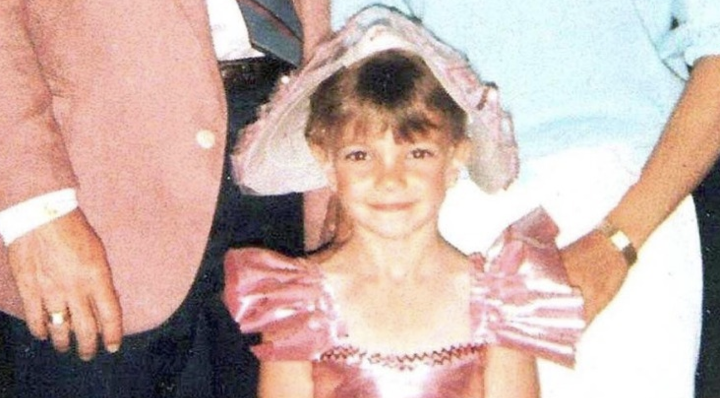 A young girl in a shiny pink dress and large matching hat smiles at the camera. Two adults, partially out of frame, stand behind her.