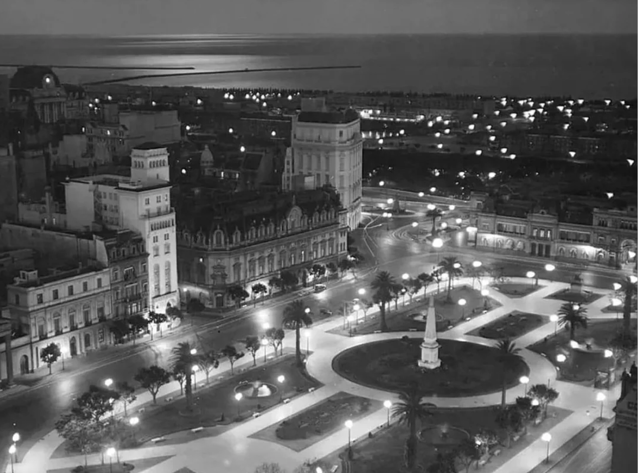 A black and white night view of a city square with an obelisk in the center, surrounded by lit pathways, palm trees, and historic buildings; the sea is visible in the background.