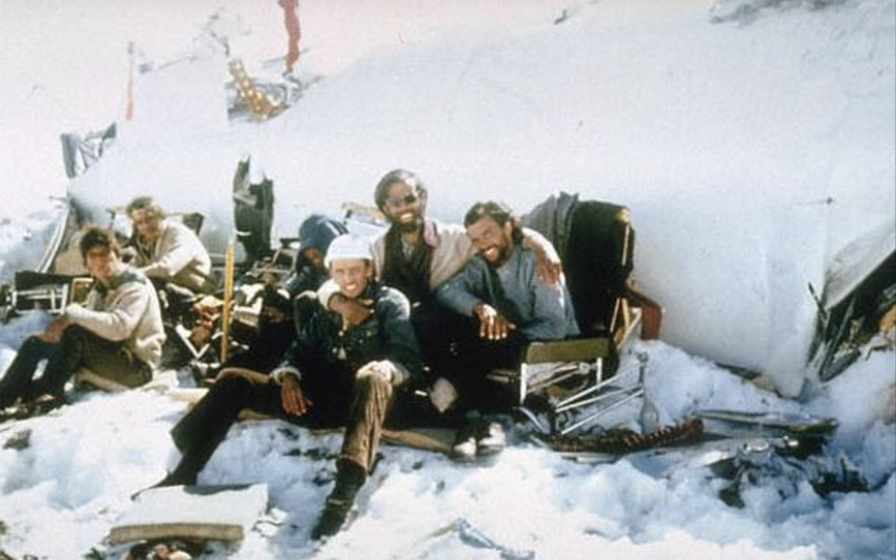 A group of men sit on snow in front of airplane wreckage, bundled in winter clothing and surrounded by scattered debris on a snowy mountainside.