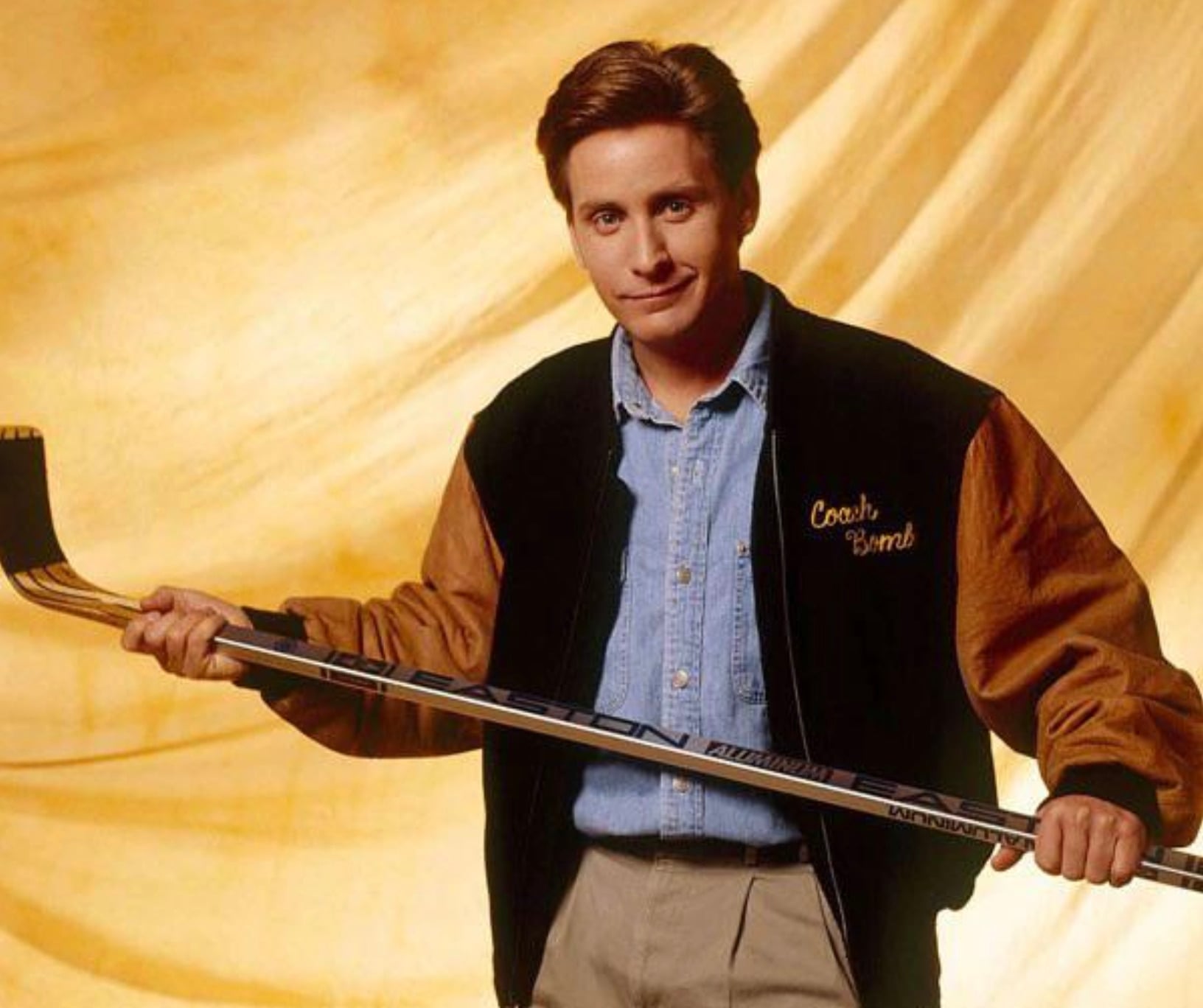 A person wearing a black and brown varsity jacket that says "Coach Bombay" holds a hockey stick and smiles against a golden yellow backdrop.