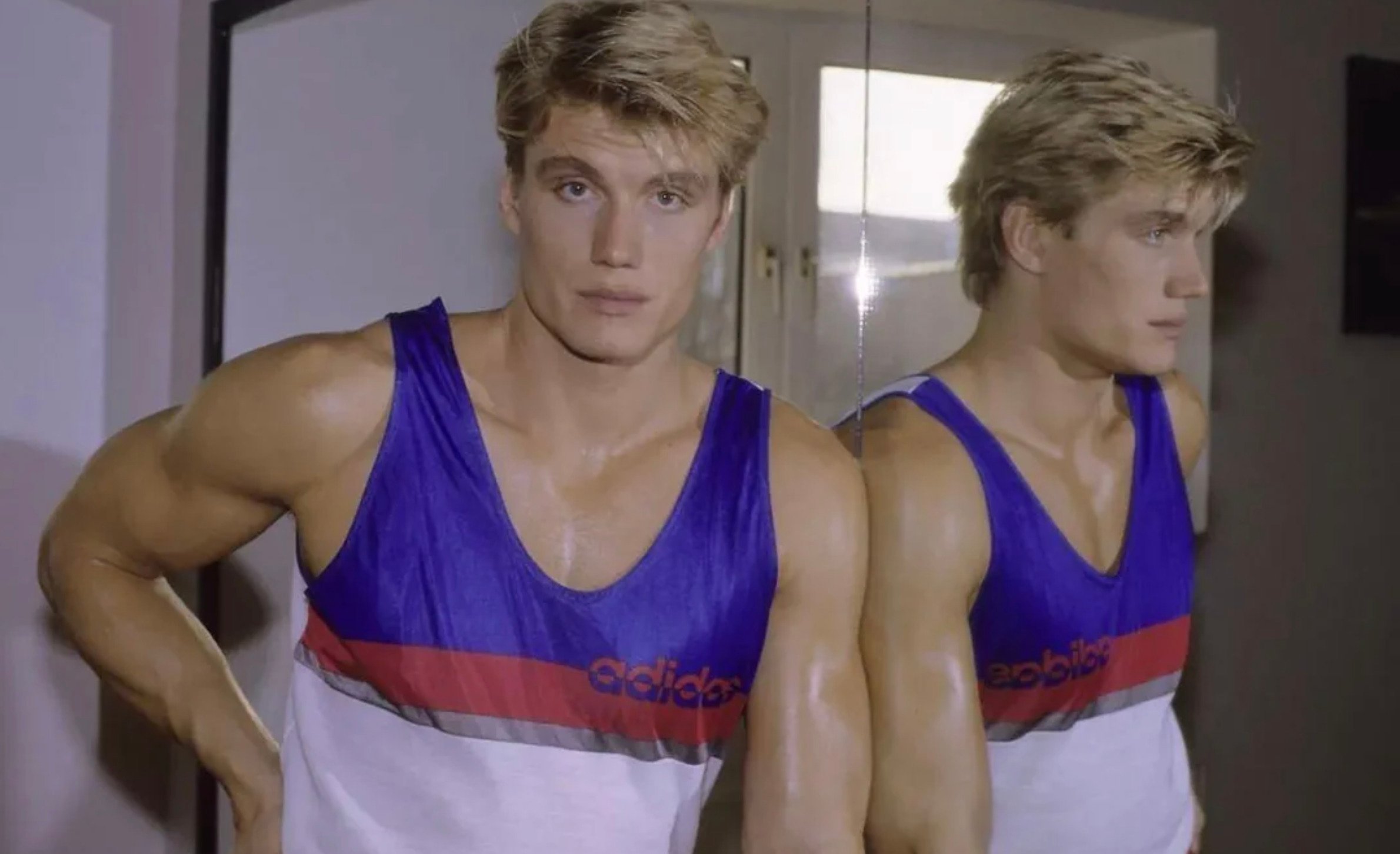 A muscular man in a sleeveless Adidas tank top stands indoors with one hand on his hip, looking at the camera. He is standing in front of a mirror reflecting his side profile.