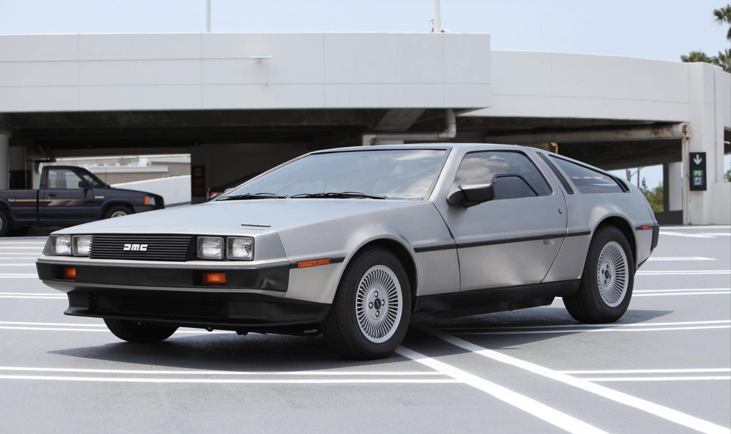 A silver DeLorean DMC-12 sports car with gull-wing doors is parked in a modern, open-air parking lot with a multi-level structure in the background.