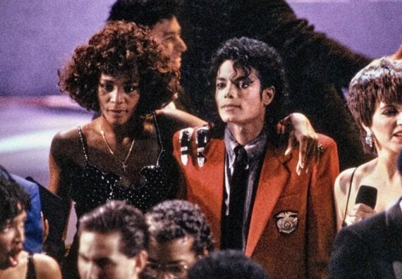 Two people stand close together at a crowded event. One wears a sparkly black dress, the other a red jacket with black details and a white shirt. Both appear relaxed, surrounded by others in evening attire.