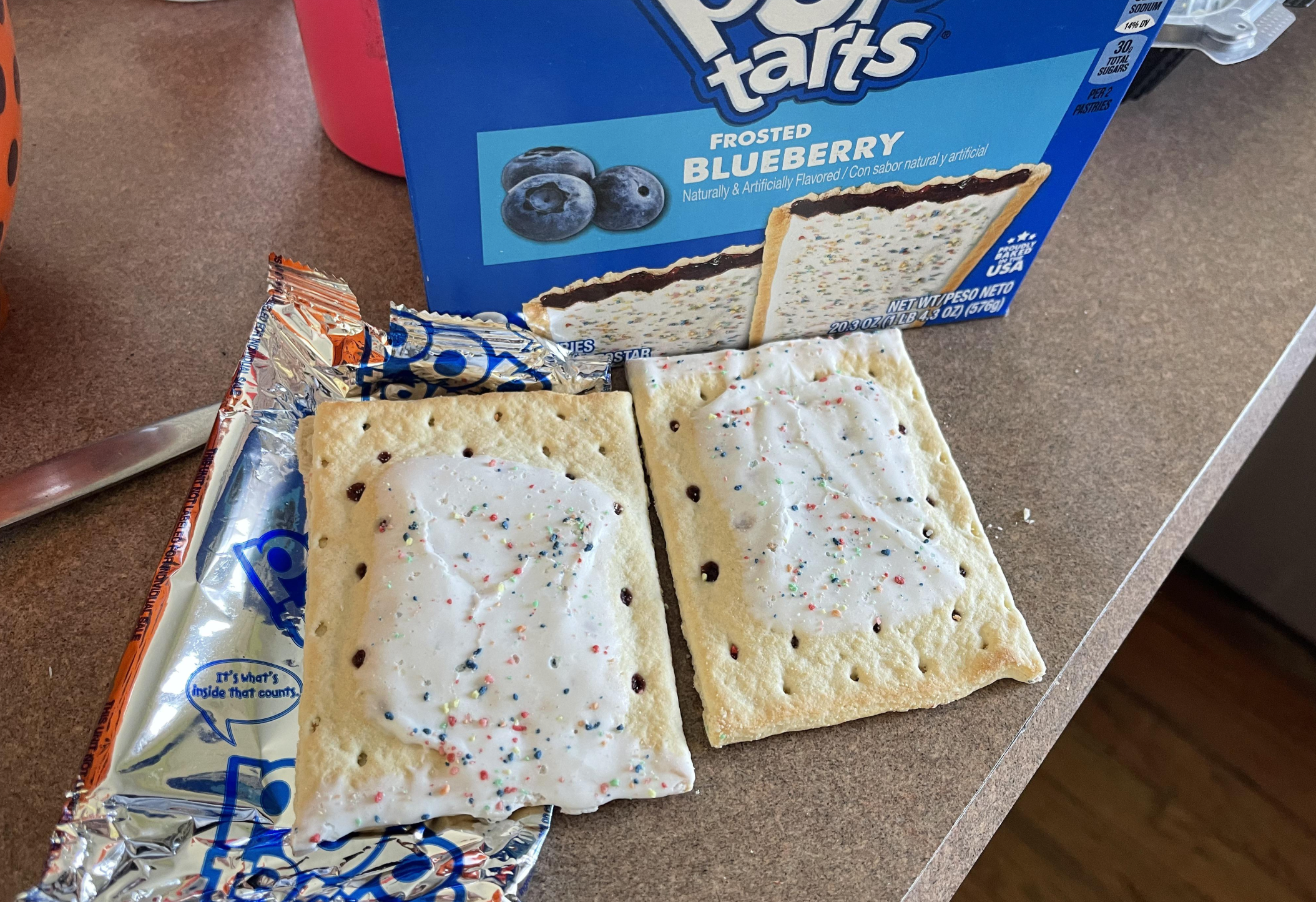 Two frosted blueberry Pop-Tarts sit on a countertop, one partially on its opened foil wrapper and both next to their blue Pop-Tarts box. The pastries have white icing with colorful sprinkles.