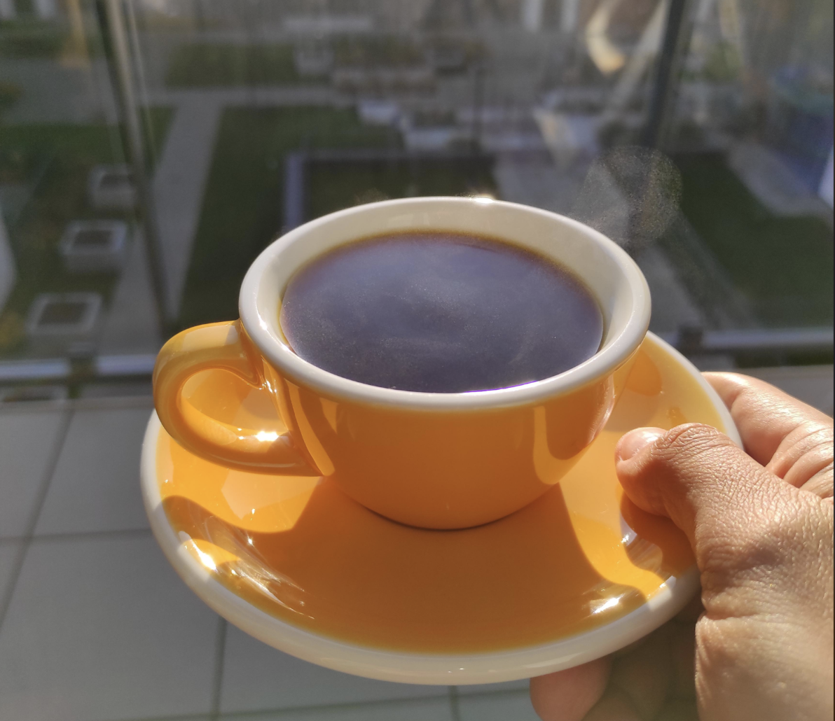 A hand holds a yellow cup and saucer filled with steaming black coffee, with sunlight shining on it and a blurred outdoor background behind glass.