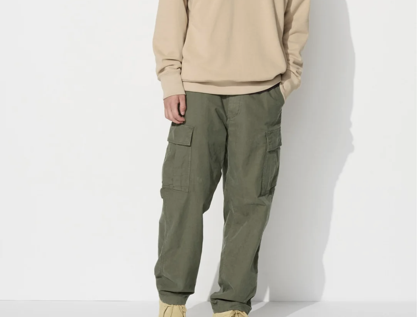 A person wearing a beige sweatshirt, olive green cargo pants with side pockets, and beige shoes stands against a white background. The person's face is not visible.