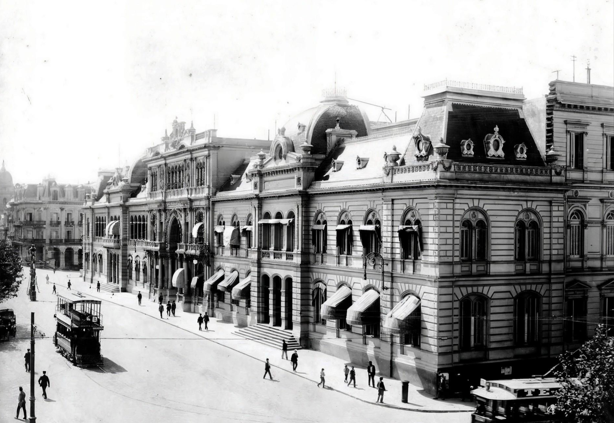 Historic black-and-white photo of a grand, ornate building with arches and domed roofs, awnings over windows, people walking on the street, and an old-fashioned tram in the foreground.