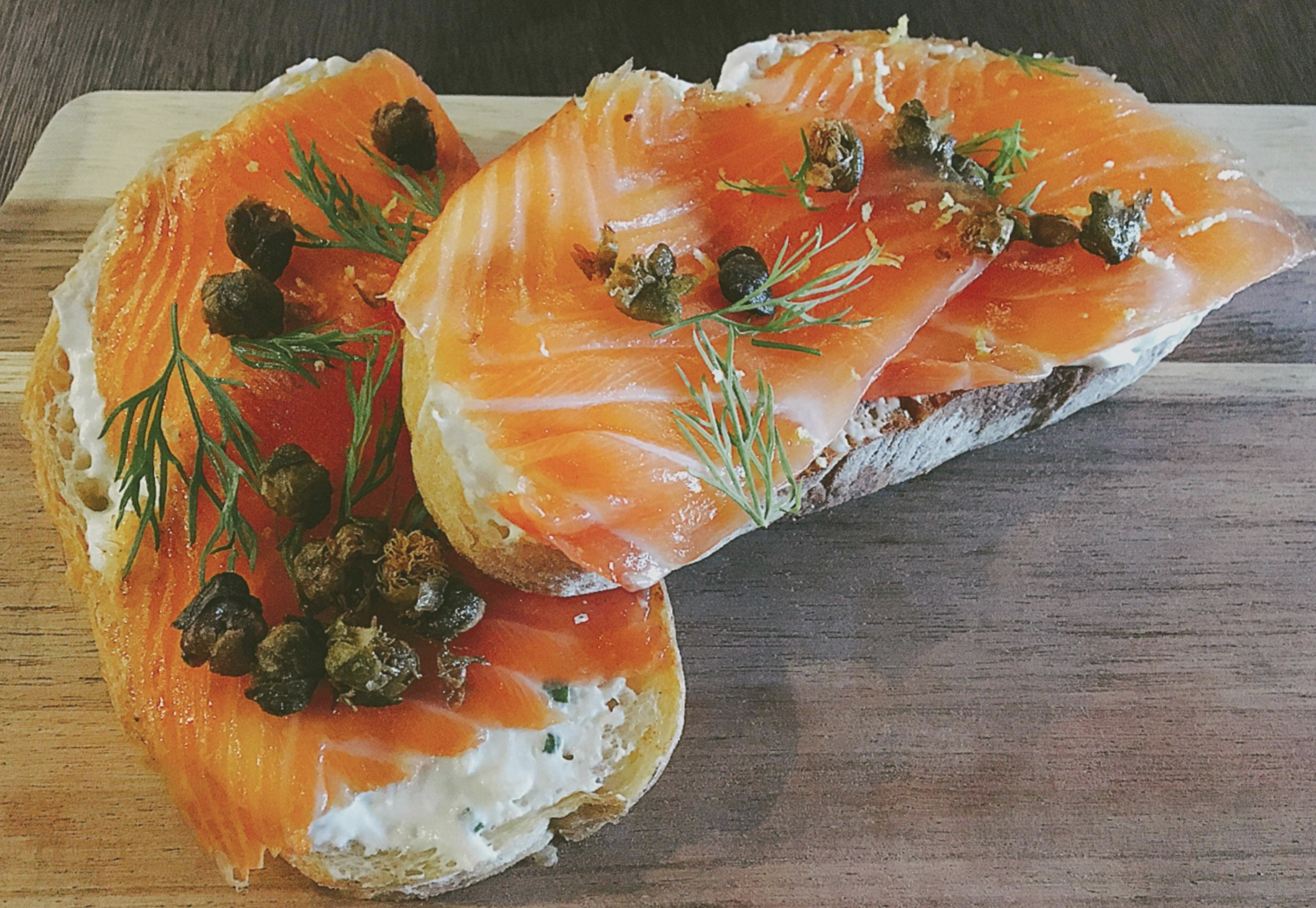 Two slices of bread topped with cream cheese, smoked salmon, capers, and fresh dill are placed on a wooden board.