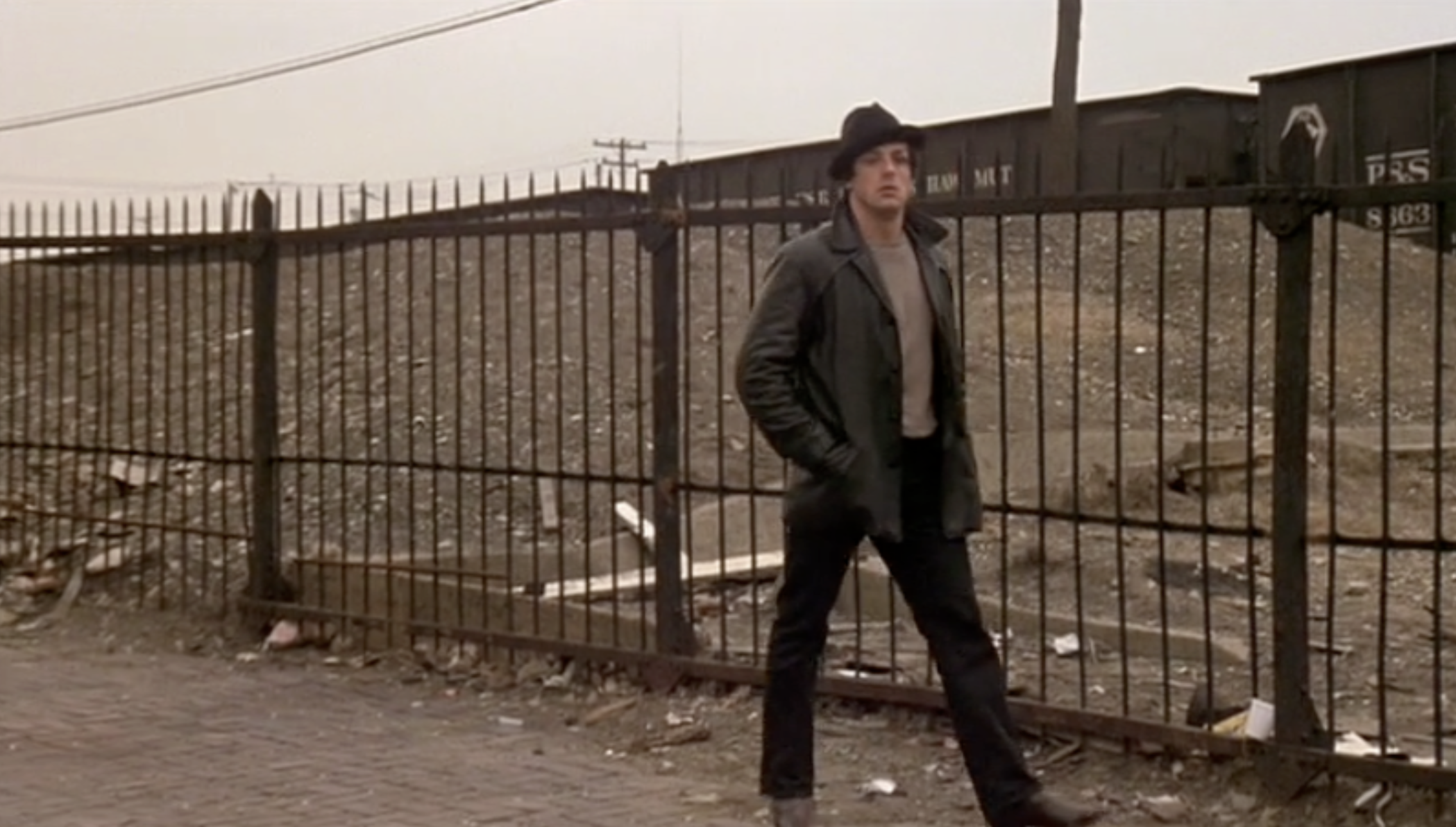 A man in a black hat and coat walks along a sidewalk beside a metal fence, with a barren, debris-strewn lot and a train car in the background under a cloudy sky.