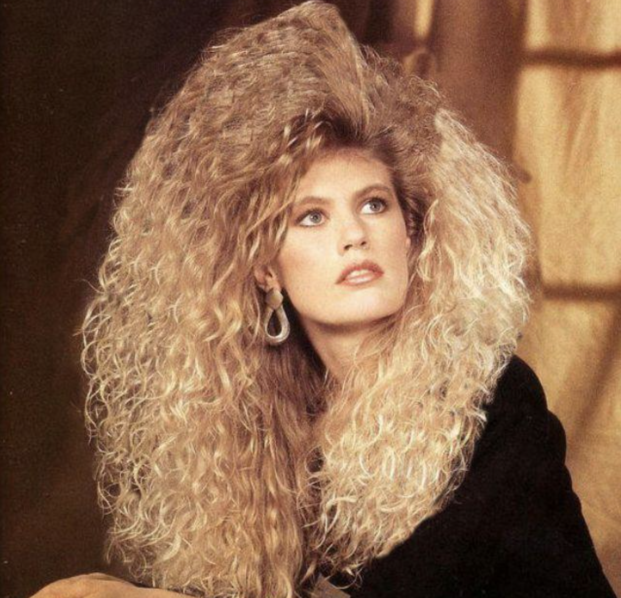 Woman with voluminous, curly blonde hair styled in an exaggerated 1980s fashion, wearing large hoop earrings and a black top, gazing upward with a soft, thoughtful expression against a warm-toned background.