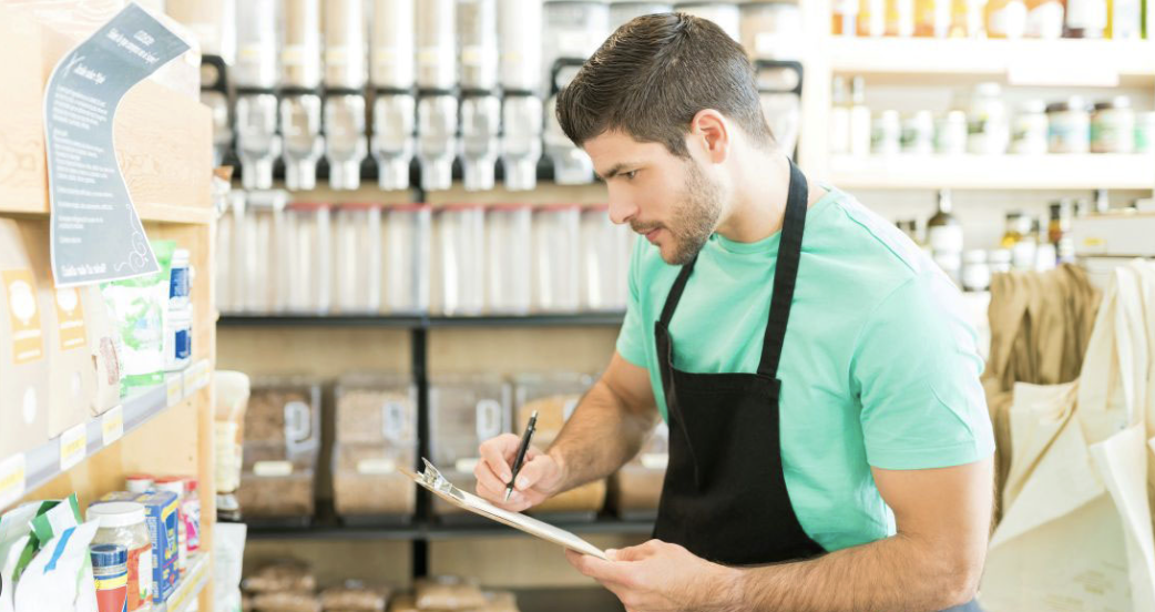 A man wearing a black apron writes on a clipboard while standing in a grocery store aisle lined with bulk food dispensers and packaged goods.