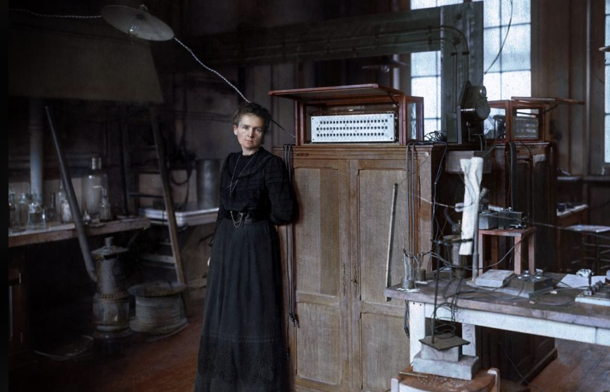 A woman in a long dark dress stands in a vintage laboratory filled with scientific equipment, wooden cabinets, and glass beakers, with large windows letting in natural light.