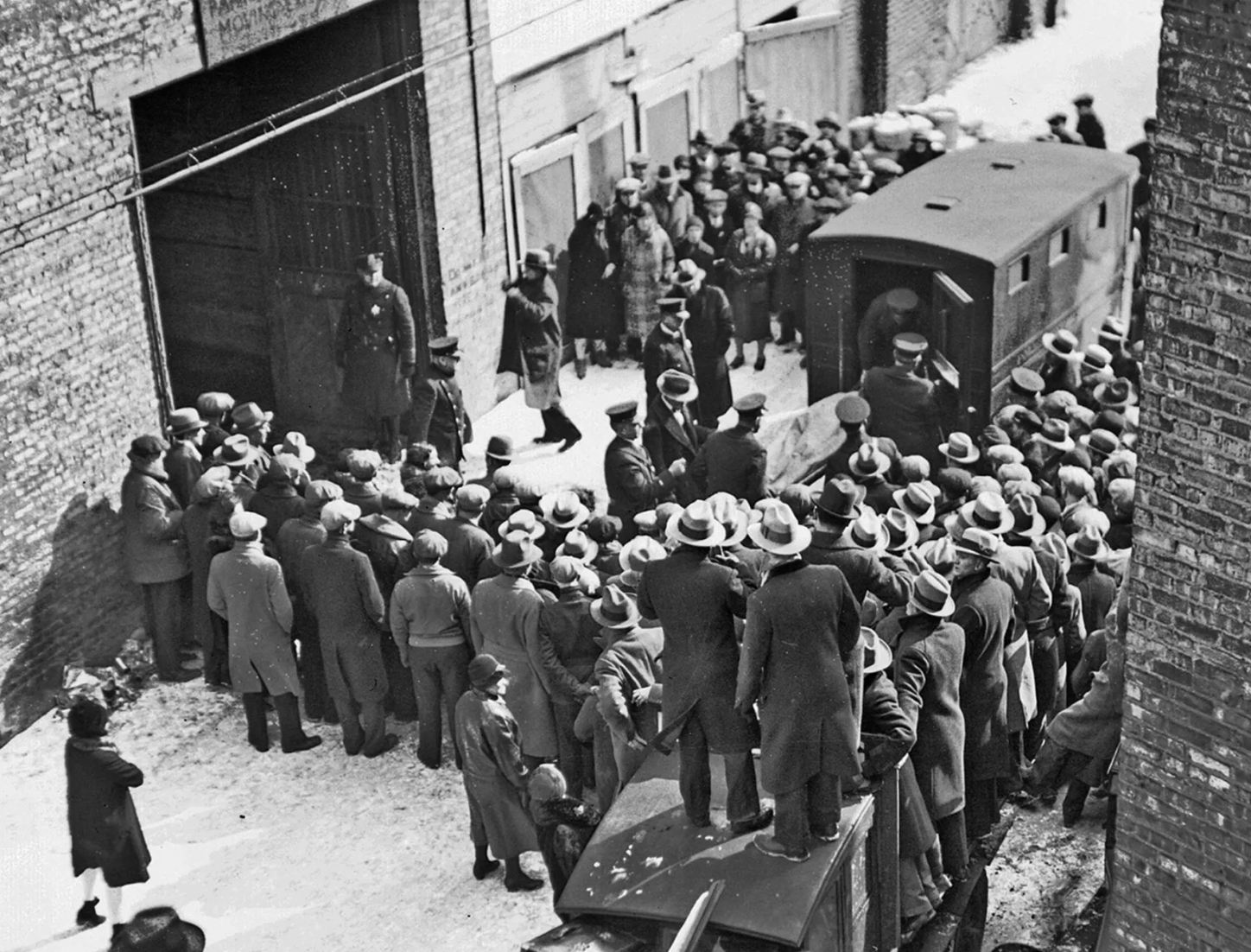 A large crowd of men in coats and hats gathers outside a building on a snowy street, watching uniformed police officers escort people into a vehicle. The scene appears tense and historic.