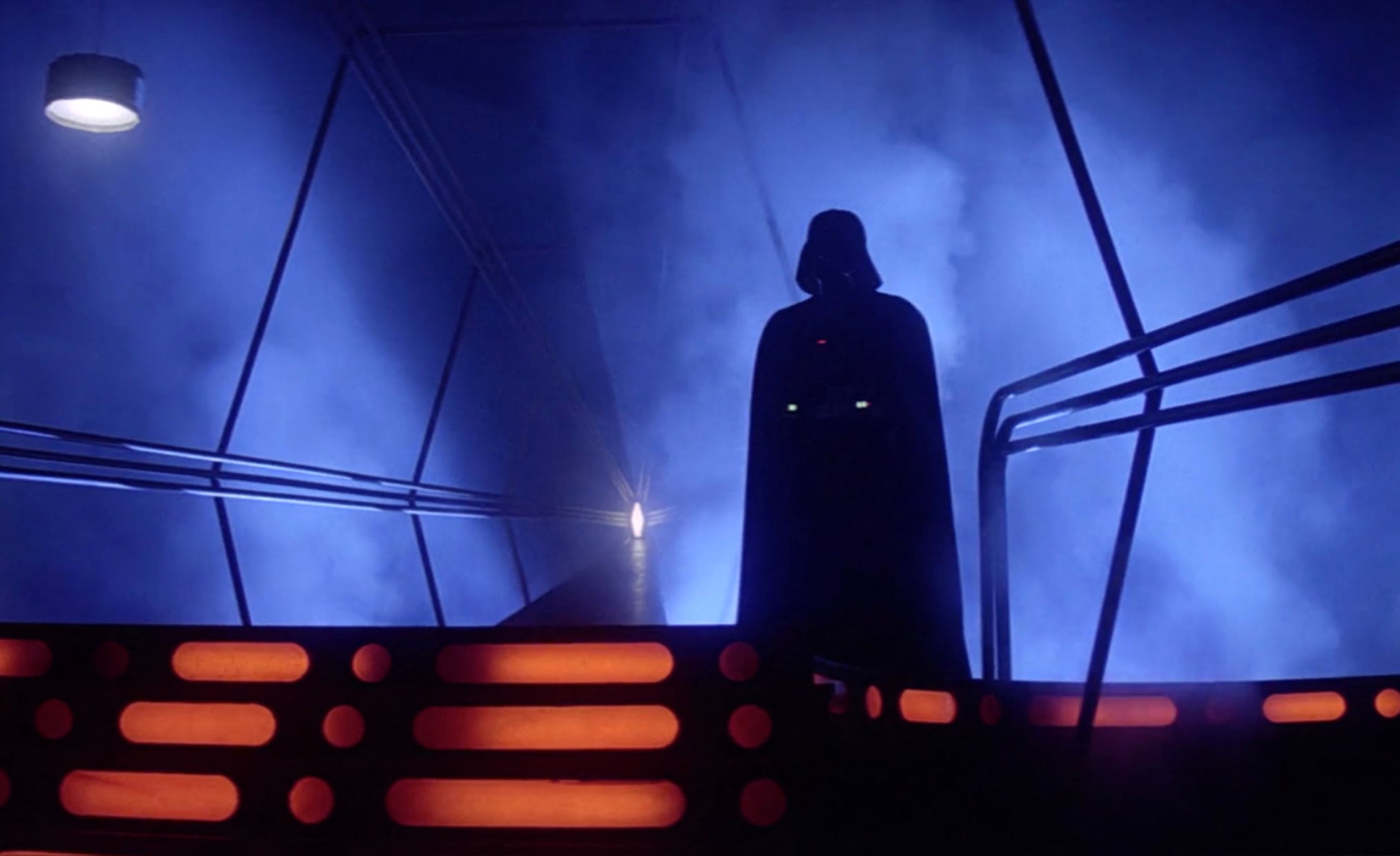 A dark figure in a cape and helmet stands on a futuristic, orange-lit platform, surrounded by metal railings and blue mist in a dimly lit, sci-fi environment.