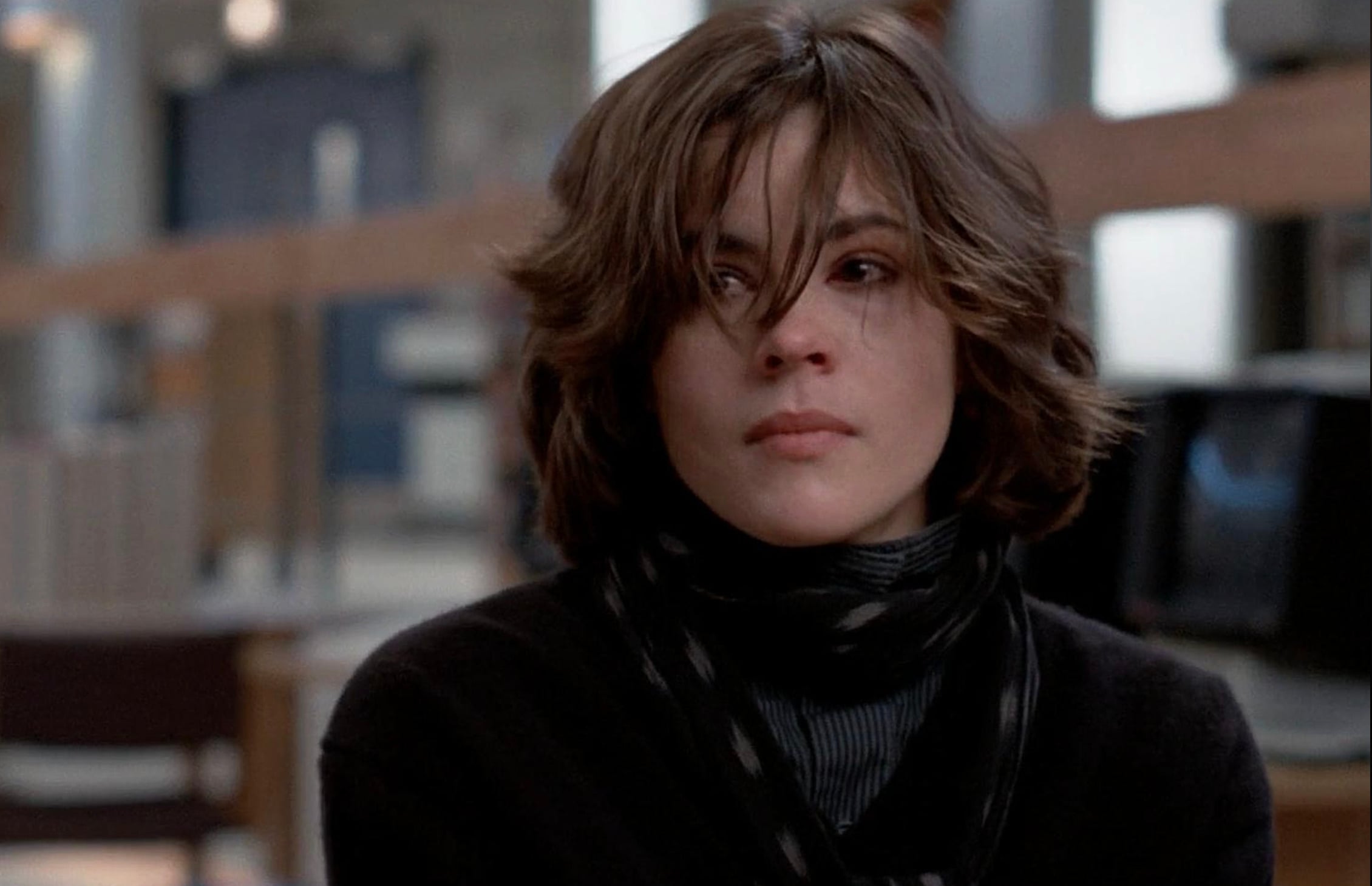 A young woman with short, tousled brown hair sits indoors, looking to the side with a serious expression. She wears a black sweater, a striped scarf, and a gray turtleneck. The background is blurred.