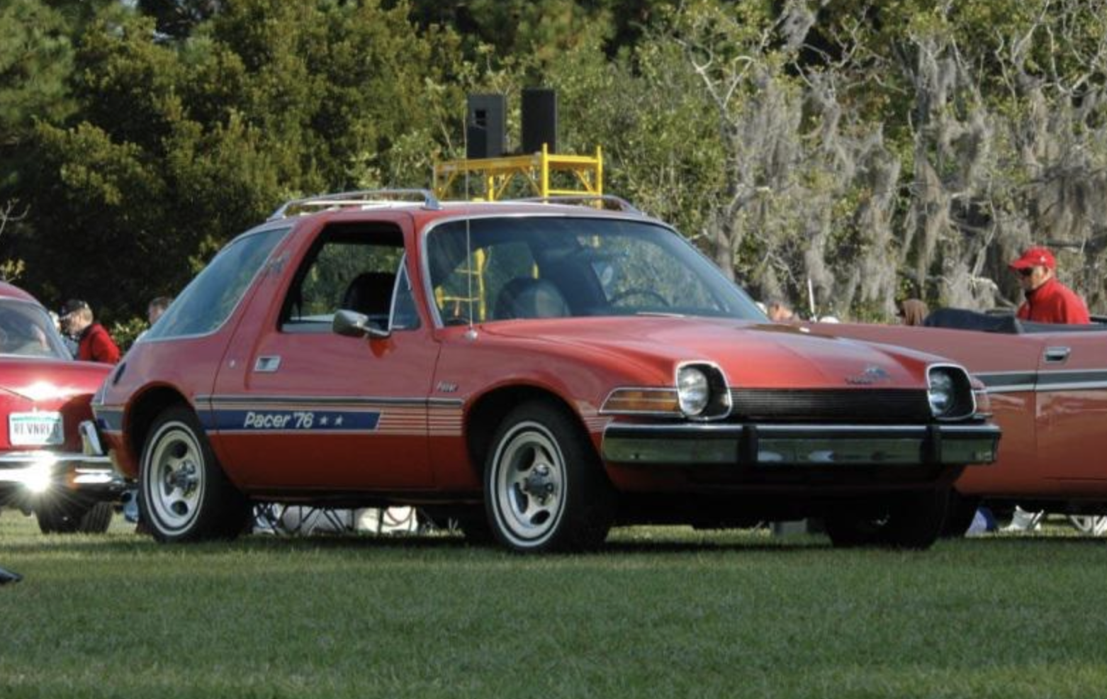 A red 1976 AMC Pacer with blue and white stripes is parked on grass at a car show, with other classic cars and people in the background.
