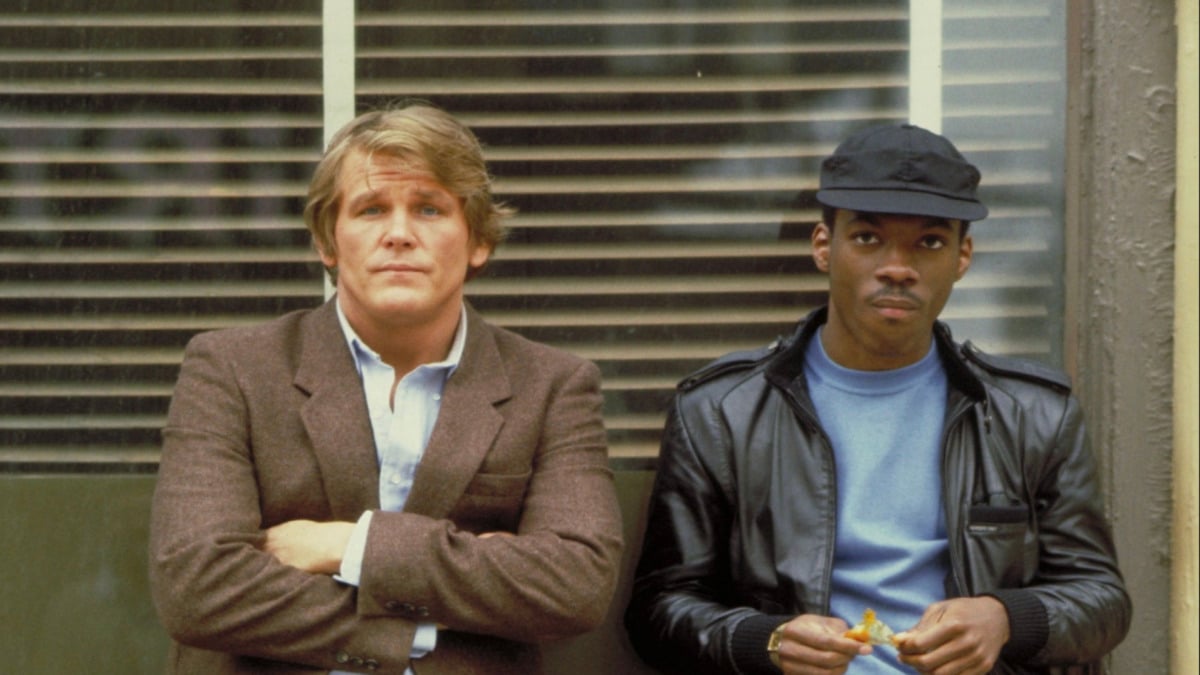 Two men stand side by side in front of a window with blinds. One wears a brown blazer with arms crossed, looking ahead; the other wears a black leather jacket and cap, holding food and looking slightly to the side.