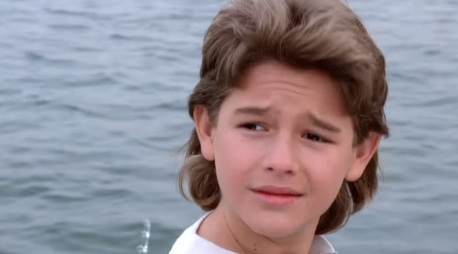 A young boy with brown, feathered hair stands in front of a body of water, looking slightly to his side with a subtle, thoughtful expression.