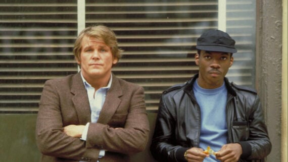 Two men stand side by side. The man on the left wears a brown blazer and looks at the camera with his arms crossed, while the man on the right wears a black leather jacket and cap, holding food in his hands. They stand in front of a window with blinds.