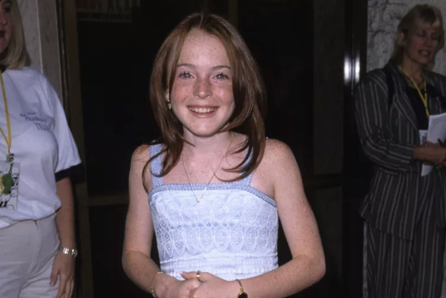 A young girl with long red hair and freckles smiles at the camera. She is wearing a light blue sleeveless dress and standing indoors, with two adults partially visible in the background.