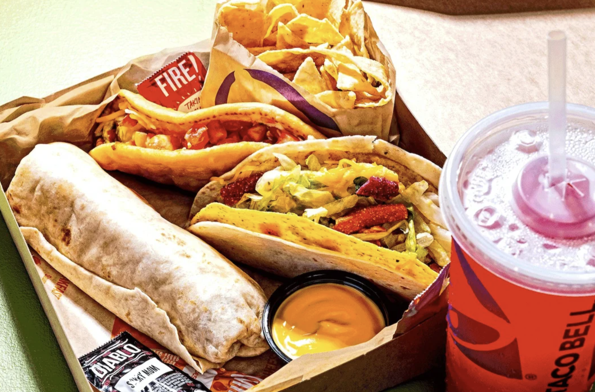 A fast food meal with a burrito, two tacos with various fillings, nacho chips with cheese sauce, hot sauce packets, and a large red soft drink in a clear cup with a straw, all in a cardboard tray.