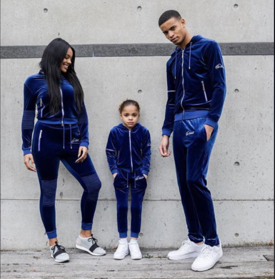 A woman, a man, and a young girl stand side by side outdoors, all wearing matching blue velour tracksuits and white sneakers, posing and smiling in front of a gray concrete wall.