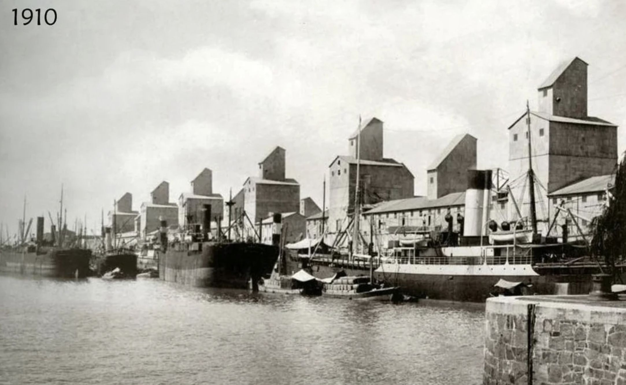 Black and white photo from 1910 showing multiple large grain elevators and docked cargo ships along a busy industrial waterfront. The water is calm, and several smaller boats are also visible.