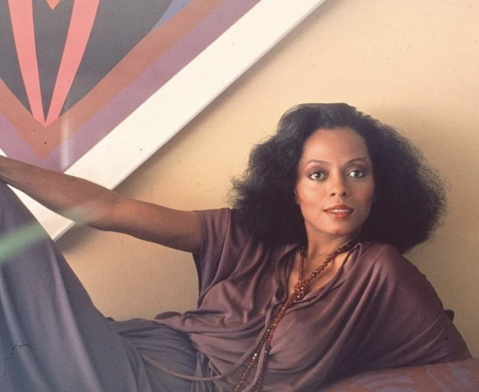 A woman with voluminous dark hair reclines on a couch, wearing a mauve outfit and long beaded necklaces, with colorful geometric art displayed on the wall behind her.