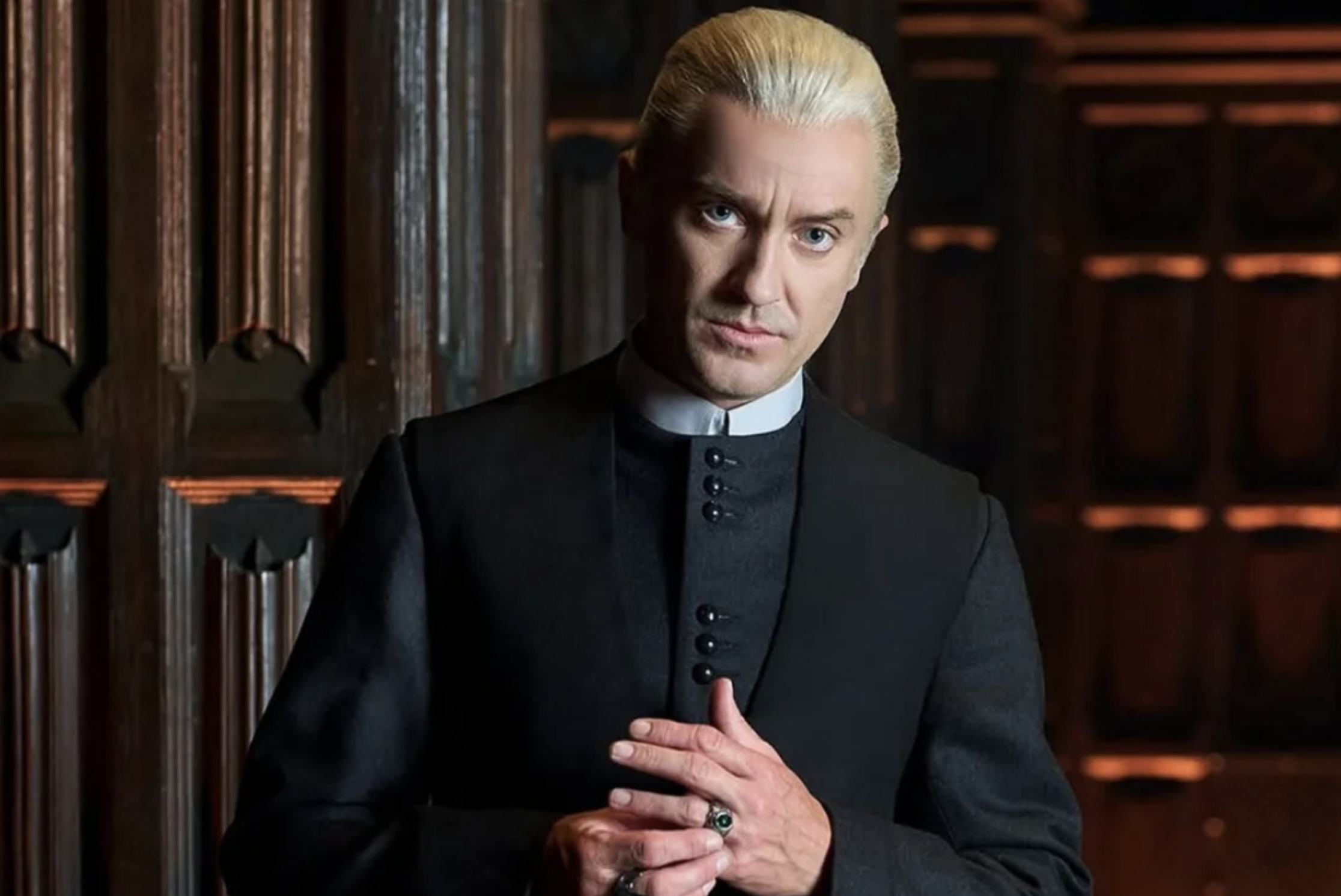 A man with slicked-back blond hair, wearing a high-collared, dark buttoned coat, stands indoors against carved wooden panels, gazing intently at the camera with his hands loosely clasped together.