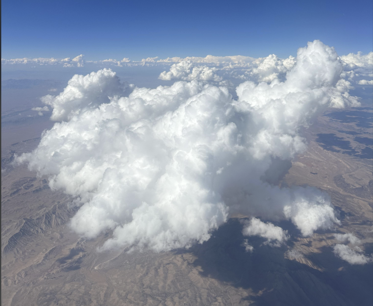 Large, fluffy white cloud floats above a dry, mountainous desert landscape, with a bright blue sky and more distant clouds stretching across the horizon.
