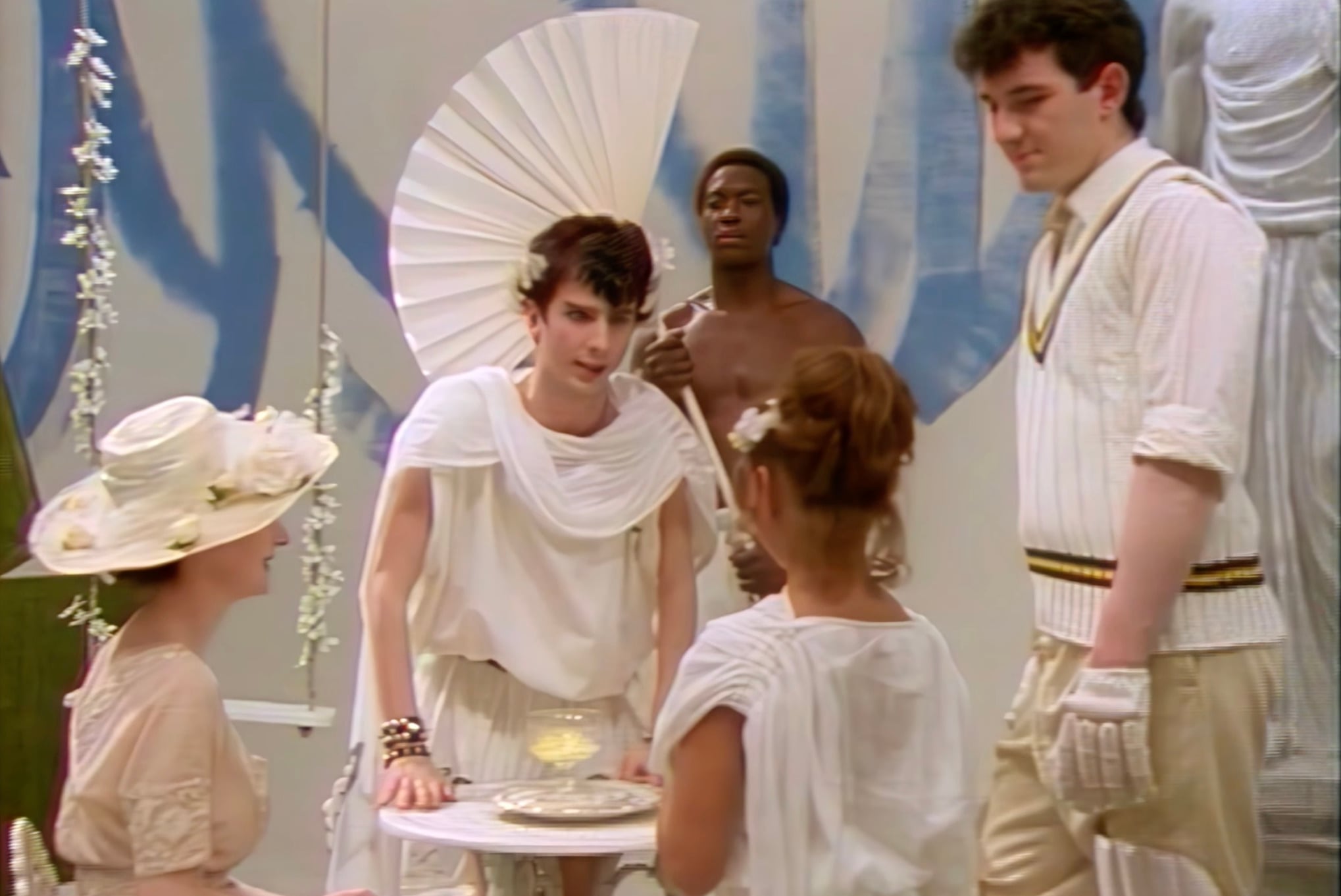 A group of people dressed in white, vintage-style clothing gather around a small table with a candle. Some wear hats and accessories; one person holds a cricket bat. The scene has a theatrical, whimsical atmosphere.