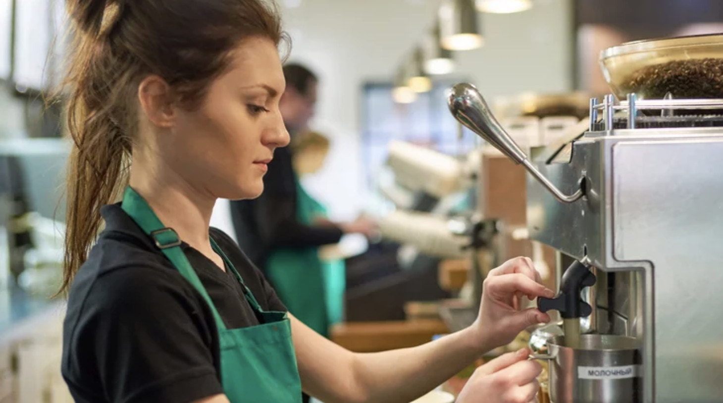 A barista in a green apron steams milk using an espresso machine in a coffee shop, focusing on her task. Another person is blurred in the background near the counter.