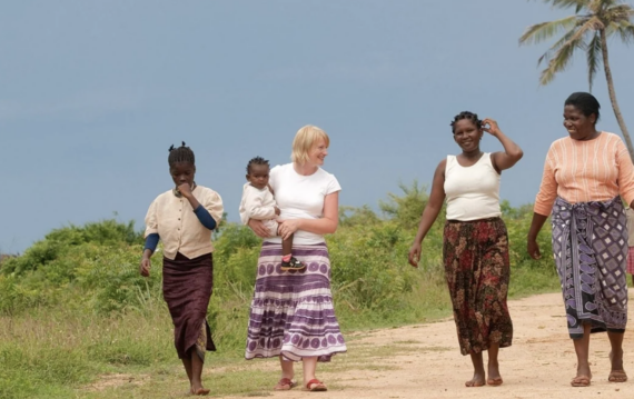 Five women walk together on a dirt path under a blue sky; one woman in the center carries a small child. They are dressed in colorful skirts and tops, and vegetation surrounds them, with palm trees in the background.
