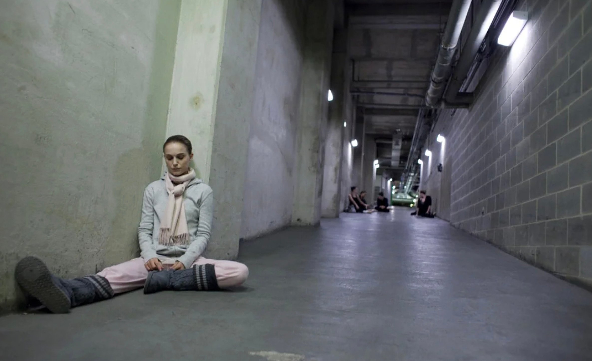 A woman in dance attire sits on the floor in a dimly lit concrete hallway, stretching with her legs bent. In the background, several other people sit or stretch along the corridor walls.