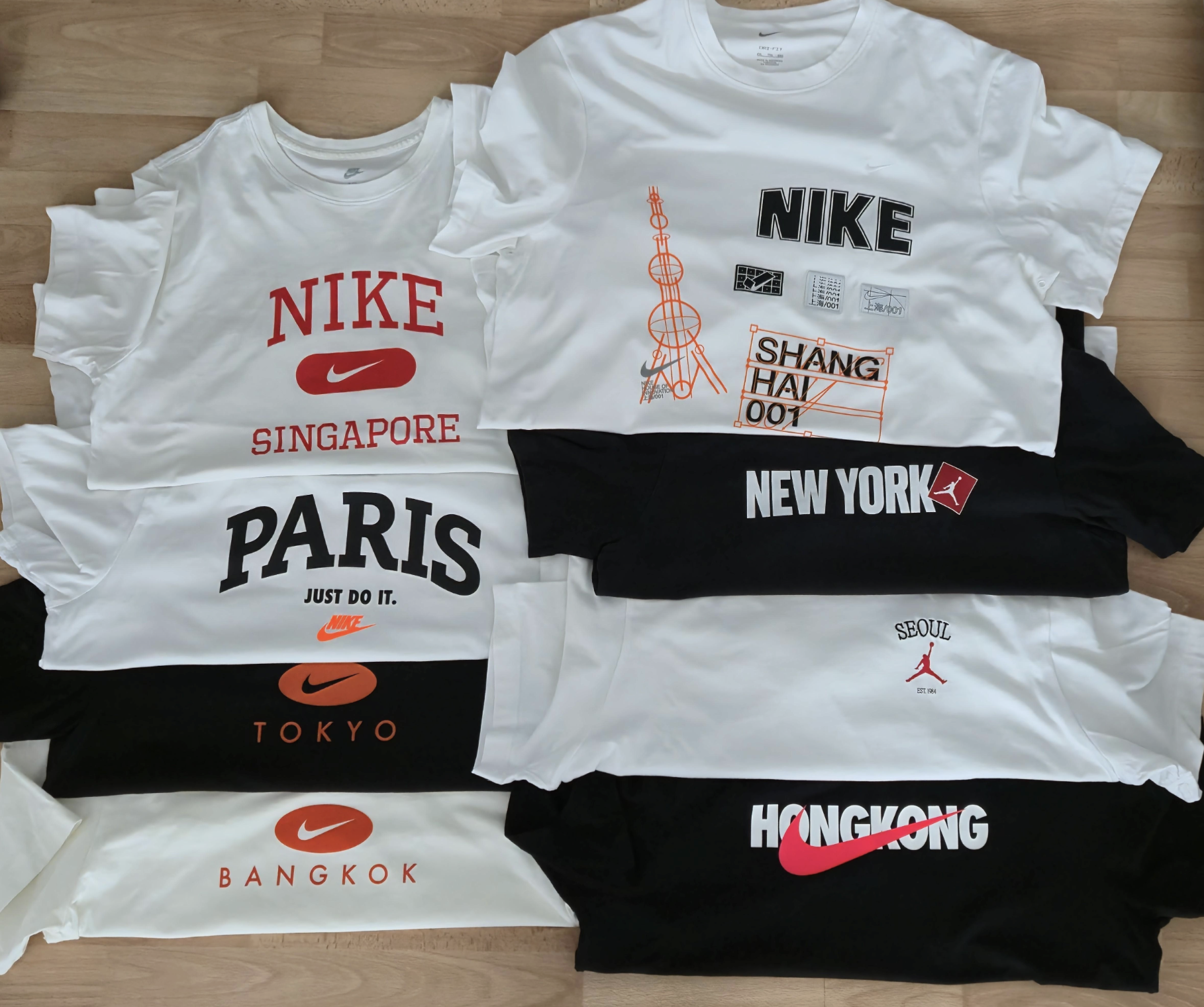A collection of folded white and black T-shirts, each displaying city names and logos such as Nike, Paris, Singapore, Shanghai, New York, Seoul, Tokyo, Bangkok, and Hong Kong, arranged on a wooden floor.