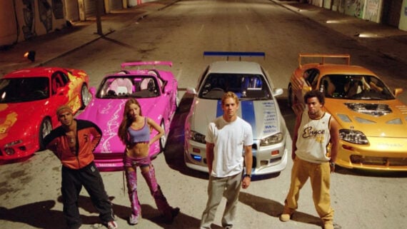 Four people stand in front of four brightly colored sports cars (red, pink, silver, and orange) on an empty city street at night, with buildings lining both sides of the road.
