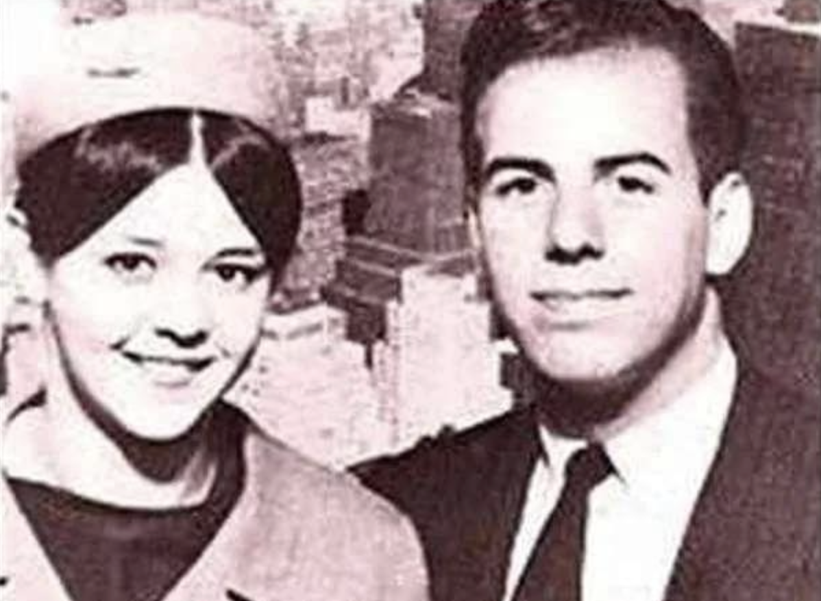 A young woman in a hat and coat smiles beside a young man in a suit and tie, both posing for a black-and-white photo with a cityscape background.