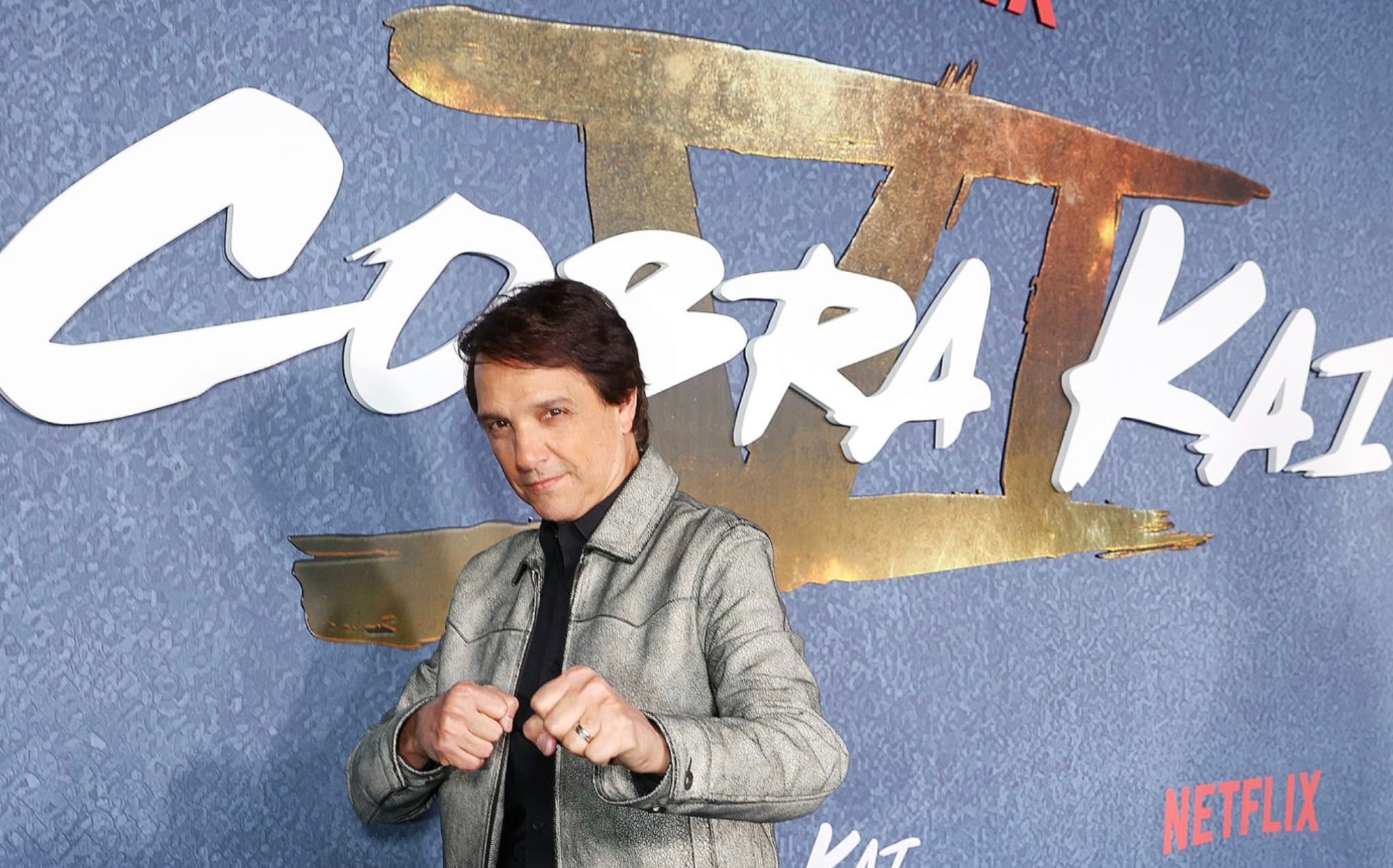 A man in a silver jacket poses with fists up in a fighting stance in front of a wall featuring the "Cobra Kai" logo and Netflix branding.