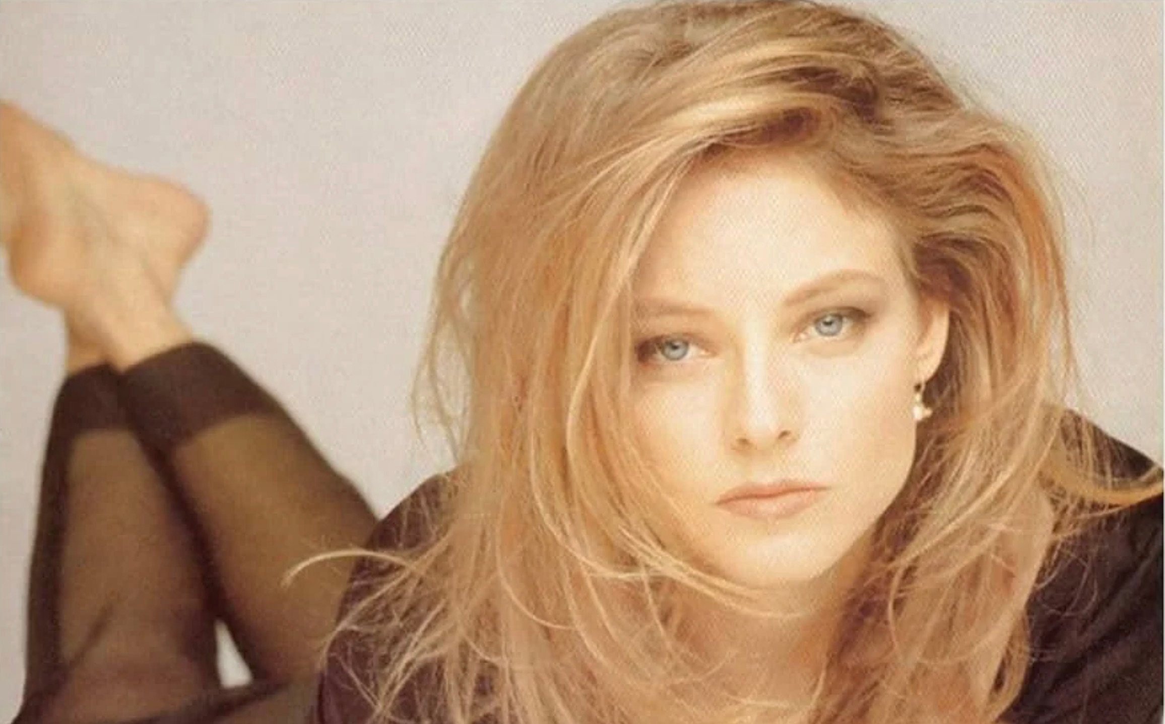 A woman with long, tousled blonde hair lies on her stomach, looking directly at the camera. She is wearing a black outfit and has a neutral expression on her face. Her feet are visible in the background.