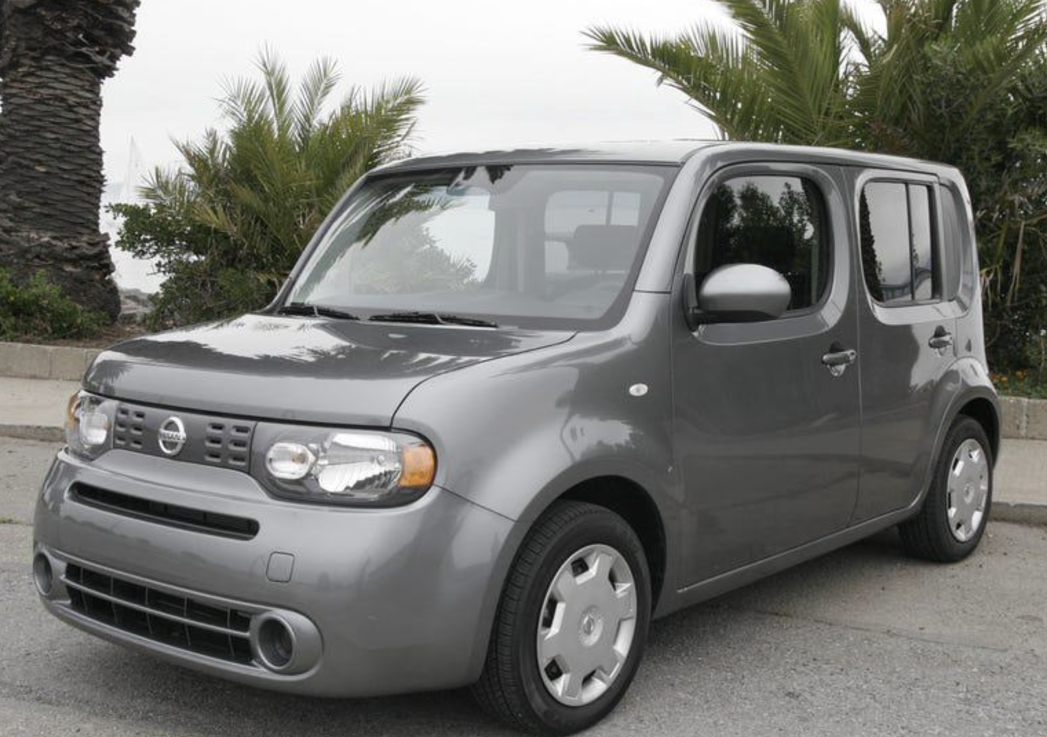 A gray Nissan Cube is parked on a street near palm trees, with a cloudy sky in the background. The car features a boxy shape and rounded edges.