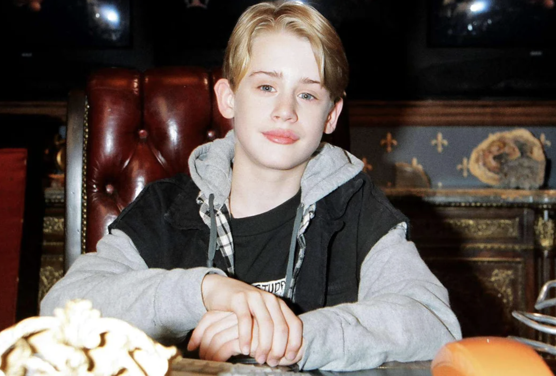 A young boy with blond hair sits at a large, ornate desk. He wears a black jacket over a gray hoodie and a T-shirt, and looks directly at the camera with a slight smile. The background is decorated with patterned wallpaper.